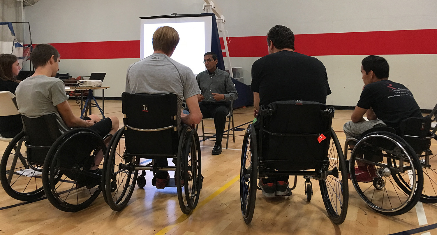 Group discussion involving two wheelchair users and a seated man in a gym.